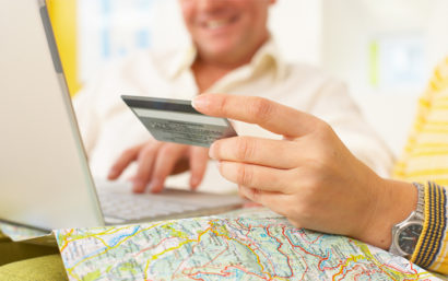 Man smiling in background while woman holds credit card in front of a laptop computer. On the table is a map.